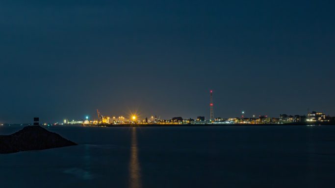 Nächtliche Küstenansicht mit beleuchteter Stadt und reflektierendem Wasser.