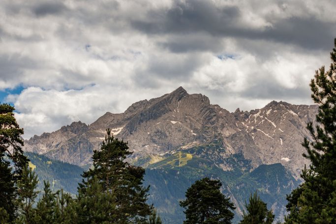Berglandschaft mit hohen Gipfeln und bewölktem Himmel, umgeben von Bäumen.