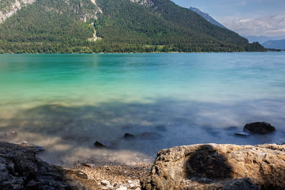 Klar blaues Wasser umgeben von Bergen und einem grünen Ufer. Steinige Küste im Vordergrund.