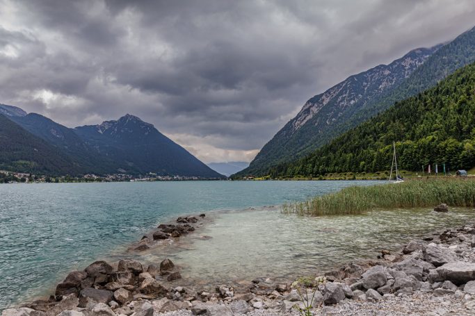 Blick auf einen ruhigen See, umgeben von hohen Bergen und bewaldeten Ufern, unter bewölktem Himmel.