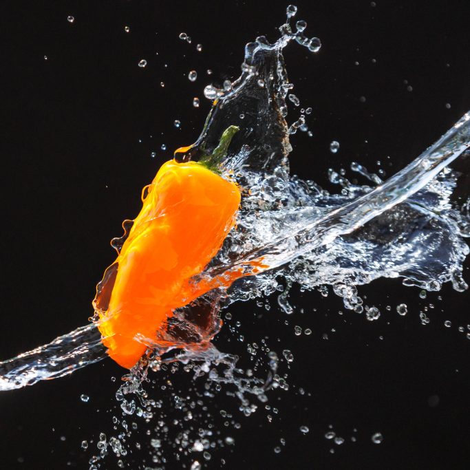 Eine orange Paprika spritzt ins Wasser vor einem schwarzen Hintergrund.