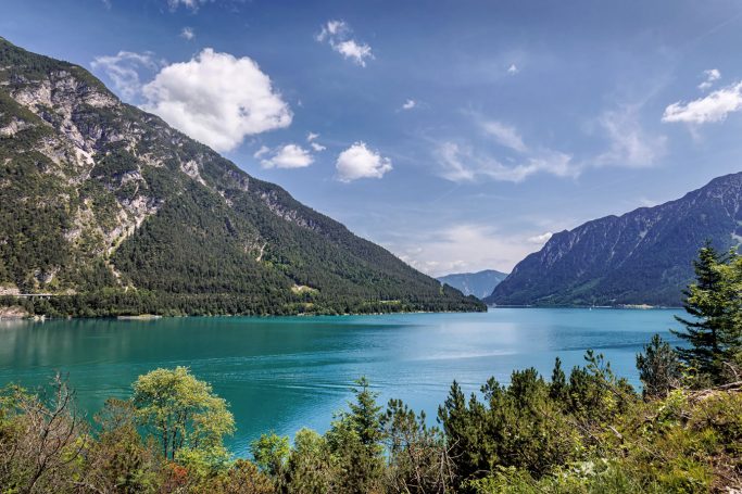 Blick auf einen klaren See umgeben von Bergen und grünem Ufer.