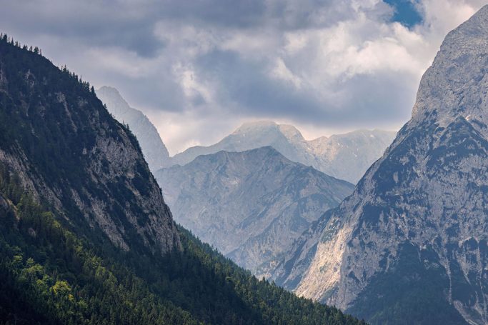 Berglandschaft mit hohen Gipfeln und bewaldeten Hängen unter bewölktem Himmel.