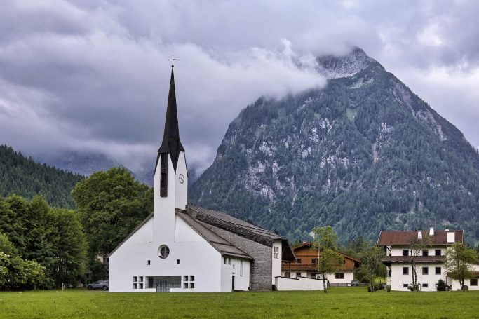 Weiße Kirche mit spitzem Turm vor einem dramatischen Bergpanorama und bewölktem Himmel.