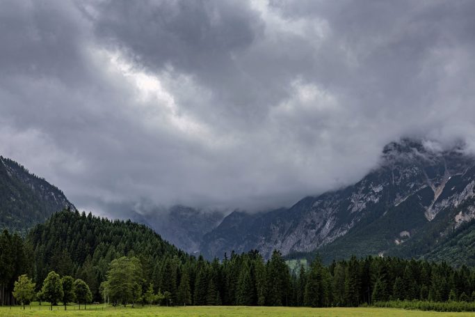 Dunkle Wolken über einem Gebirgszug, umgeben von grünen Wäldern und einer Wiese.